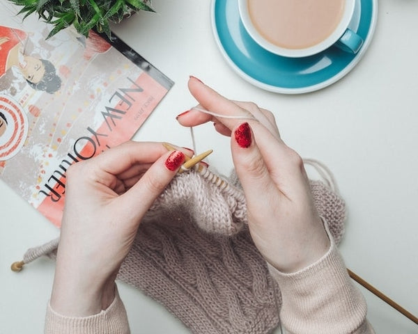 Una persona che indossa scintillanti unghie finte rosse fatte a mano mentre lavora a maglia con una rivista e una tazza di caffè vicina