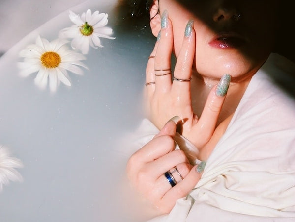 A person's hands adorned with shimmering fake nails, partially submerged in water alongside floating daisy flowers