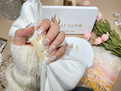 A hand displaying elegant fake nails with a clear base, adorned with gold foil details and pink accents, presented against a backdrop featuring the LTGlow brand box and a floral arrangement, embodying sophistication and modern nail artistry.