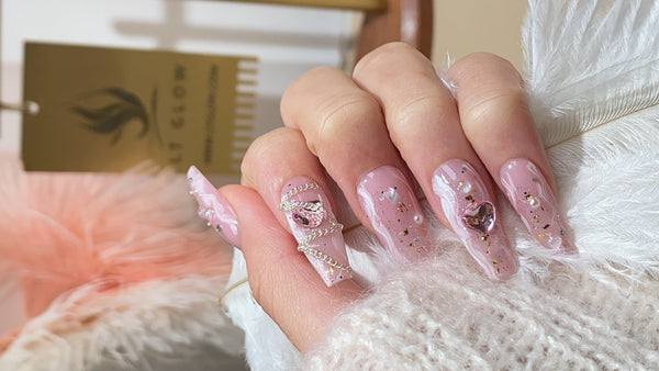 Elegant acrylic nail design featuring pink tones, intricate sparkles, and decorative gem embellishments, showcased against a fluffy backdrop