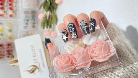 A hand presents a set of luxurious handcrafted false nails with a navy and gold design, featuring delicate floral patterns and sparkling rhinestones, poised over a clear container of soft pink artificial roses, with nail art supplies subtly visible in the background