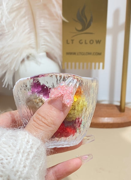 A hand adorned with handcrafted false nails featuring a colorful glitter design, holding a glass bowl, with the LT GLOW logo in the background.