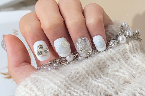 Close-up of a hand showcasing white handmade false nails with delicate floral designs and glitter accents, draped over a cozy knitted fabric with a pearl bracelet