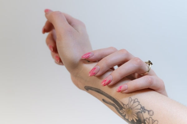 Hand showcasing handcraft fake nails in a vibrant pink shade with a floral tattoo design on the forearm.