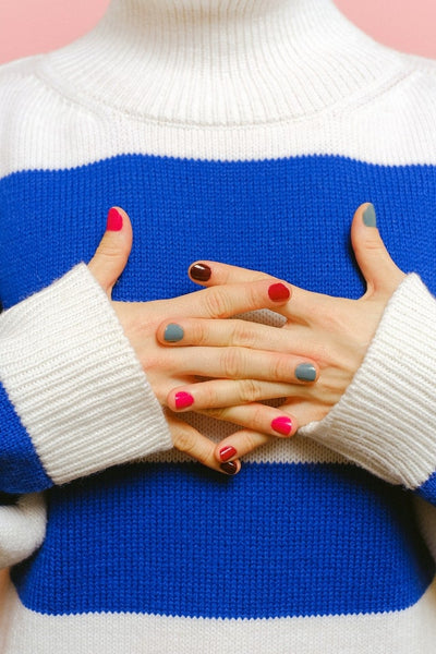 Hands crossed over a blue and white striped sweater, showcasing an array of false nails painted in varying shades of red, pink, and teal