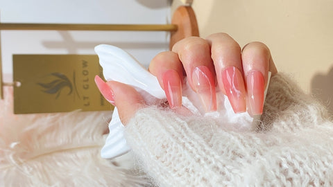 A hand features long, stiletto-shaped false nails with a gradient of soft pink to clear tips, held against a cozy background that includes the LTGlow brand identity, showcasing a modern and chic nail aesthetic