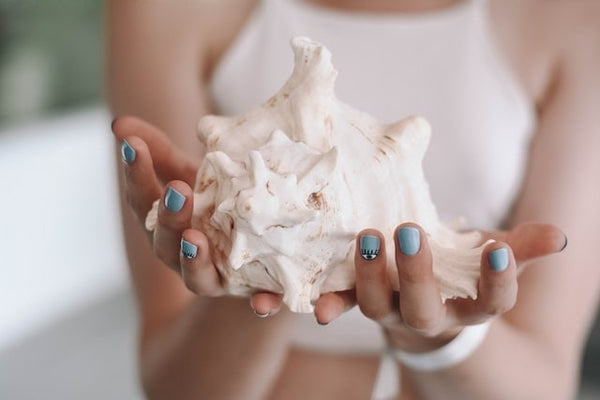 Une personne présentant des faux ongles à coller faits main bleu océan avec un design délicat, tout en tenant un grand coquillage blanc