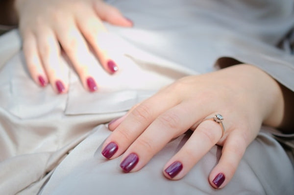 Elegant hands displaying deep burgundy handcraft false nails, accentuated by a delicate ring, resting on soft white fabric