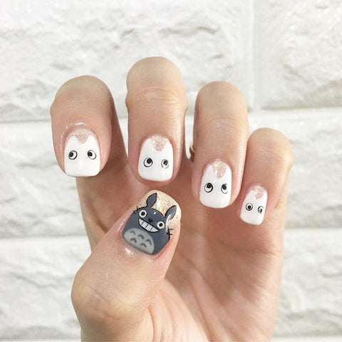 A playful set of handcrafted fake nails featuring whimsical characters with expressive eyes on a shimmering nude base, and one accent nail artfully depicting a beloved animated creature in grey and blue tones