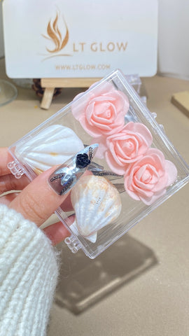 An elegantly manicured hand holds a clear box showcasing acrylic nails, one with a sophisticated navy blue design with a large gemstone, and others adorned with white and gold marble effects, all against a backdrop of delicate pink roses and the minimalist branding of "LT GLOW."