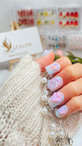 A hand showcasing shimmering opalescent false nails with delicate heart and cloud designs, complemented by a pearl bracelet and the LTGlow brand packaging in the background, encapsulating a dreamy and romantic aesthetic.