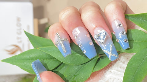 Elegant handcrafted false nails in pastel blue adorned with silver accents, rhinestones, and delicate chains, presented against a backdrop of fresh green leaves, showcasing a luxurious and creative nail art design