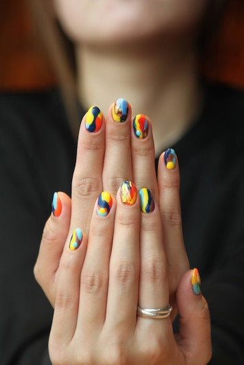 Hands showcasing vibrant handcraft false nails painted with multi-colored swirl patterns. The nails are a vivid blend of blue, yellow, and red tones