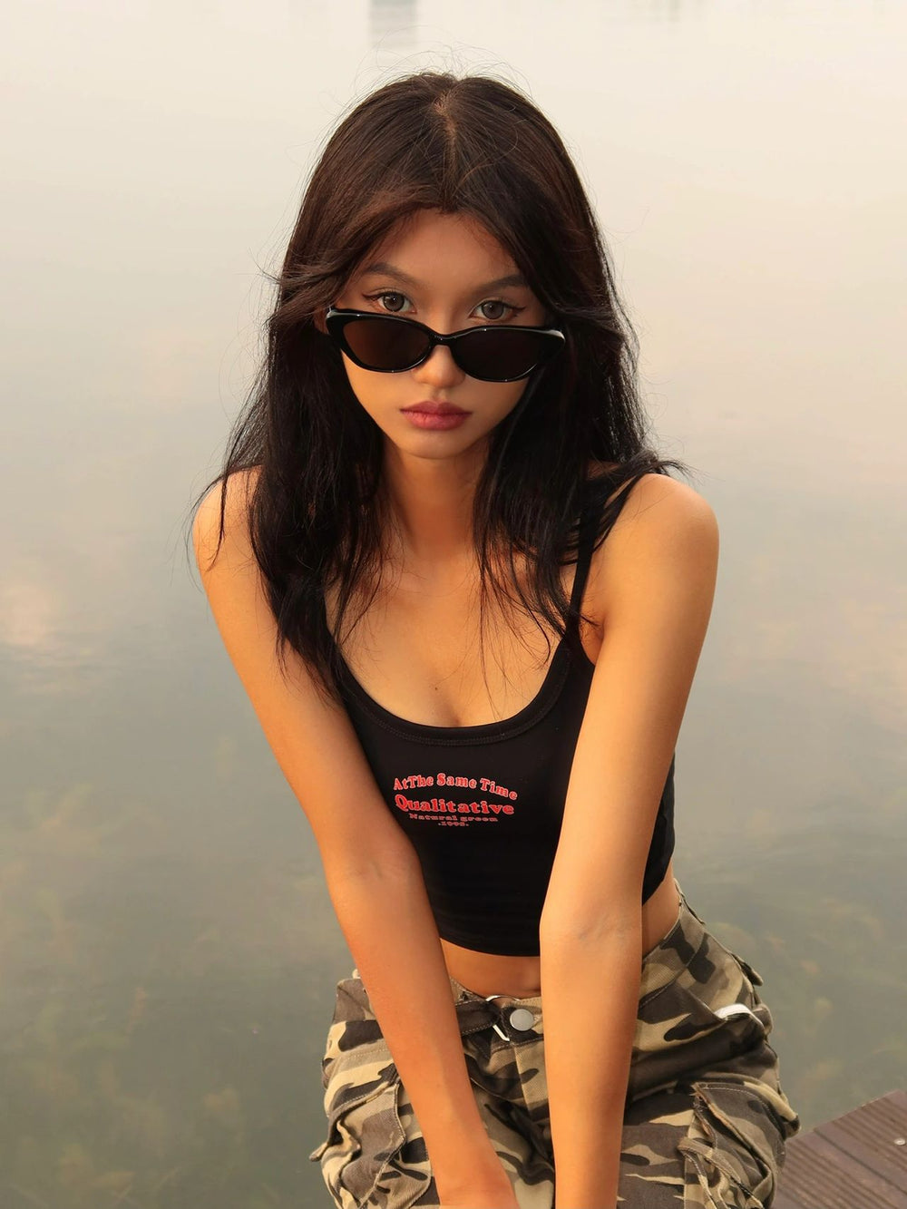 A glamorous lady, donning stylish sunglasses, gracefully perches on a rock beside the serene waters, exuding an air of elegance and tranquility.