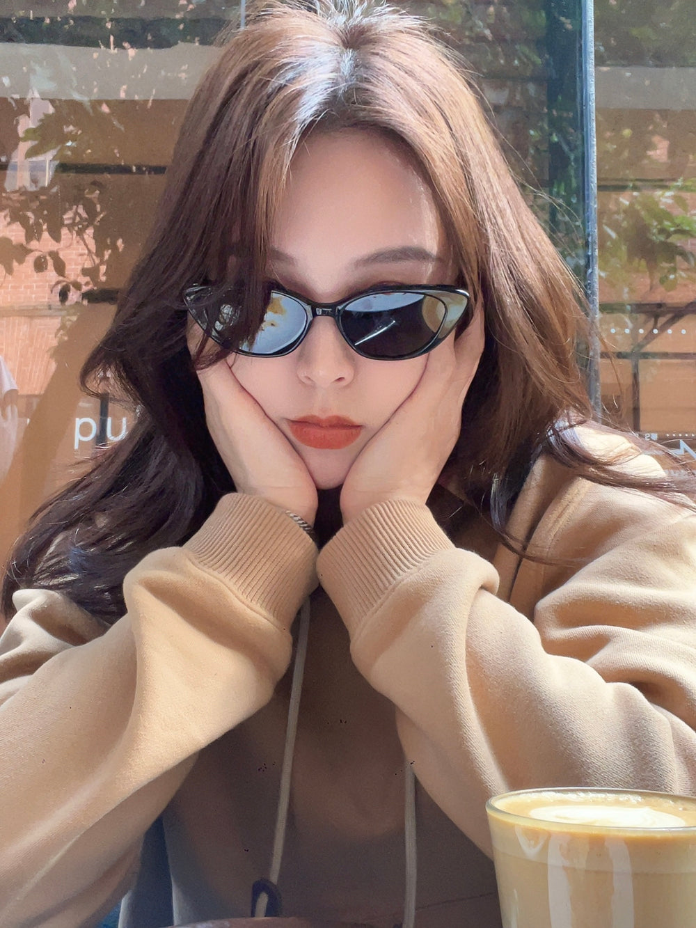 An elegant woman, adorned with fashionable sunglasses, sits at a table, her head gracefully supported by her hands.