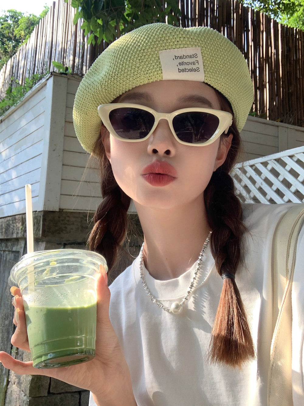 An elegant woman, adorned with a trendy hat and fashionable sunglasses, gracefully clutches a vibrant green drink.