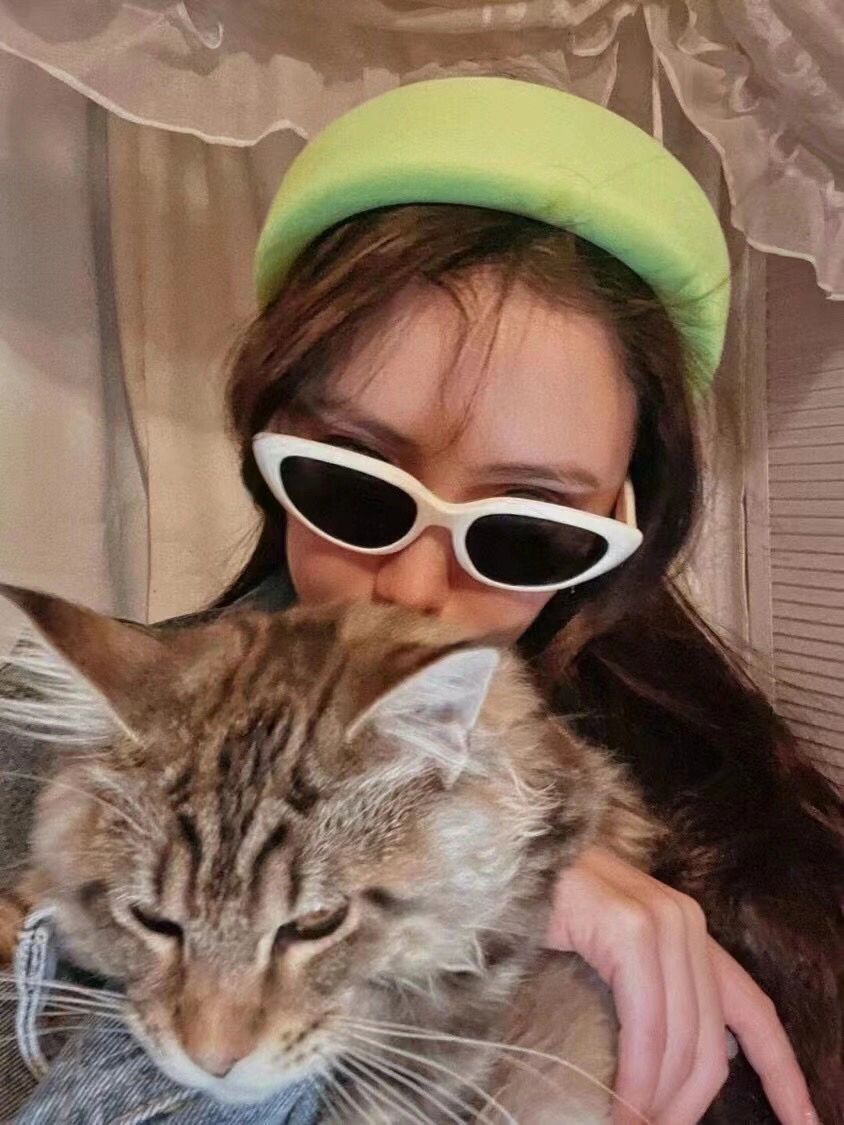 An elegant woman, adorned with a fashionable green hat and trendy sunglasses, tenderly embraces a charming cat.