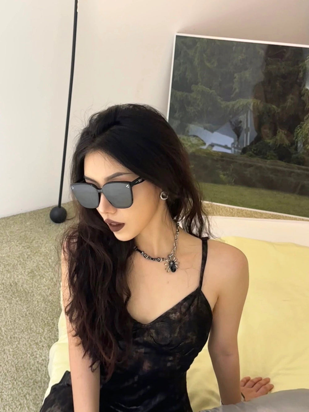 With an air of refined charm, a woman in a chic black dress sits gracefully on a luxurious bed, epitomizing sophistication and grace with her fashionable sunglasses.