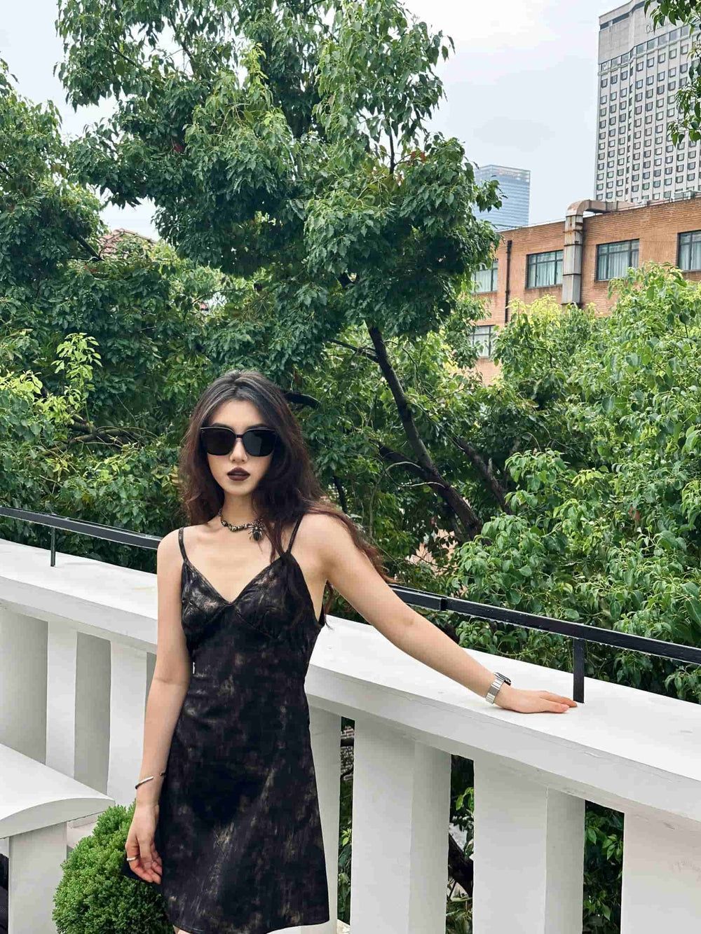 A stylish woman in a black dress and trendy sunglasses striking a pose on a balcony.