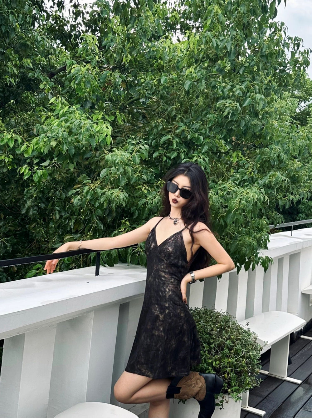 Radiating sophistication, a woman donning a chic black dress and trendy sunglasses strikes a pose on a sun-kissed deck.