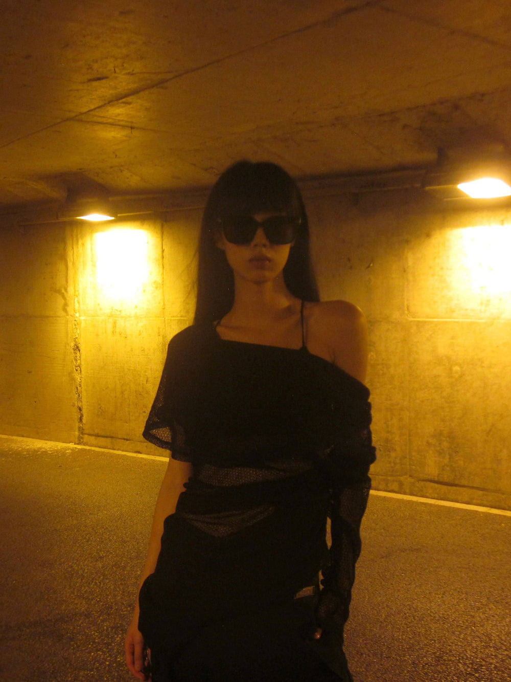 In the vast emptiness of a parking garage, a woman donning a sleek black dress commands attention with her graceful presence with her stylish sunglasses.