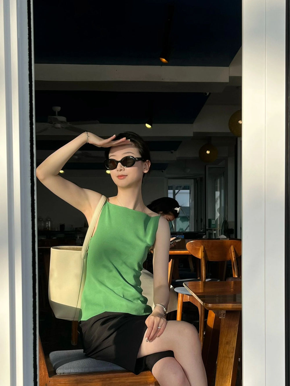 poised woman dressed in a trendy green shirt and a sleek black skirt, seated gracefully on a stylish chair with her Korean fashionable sunglasses.