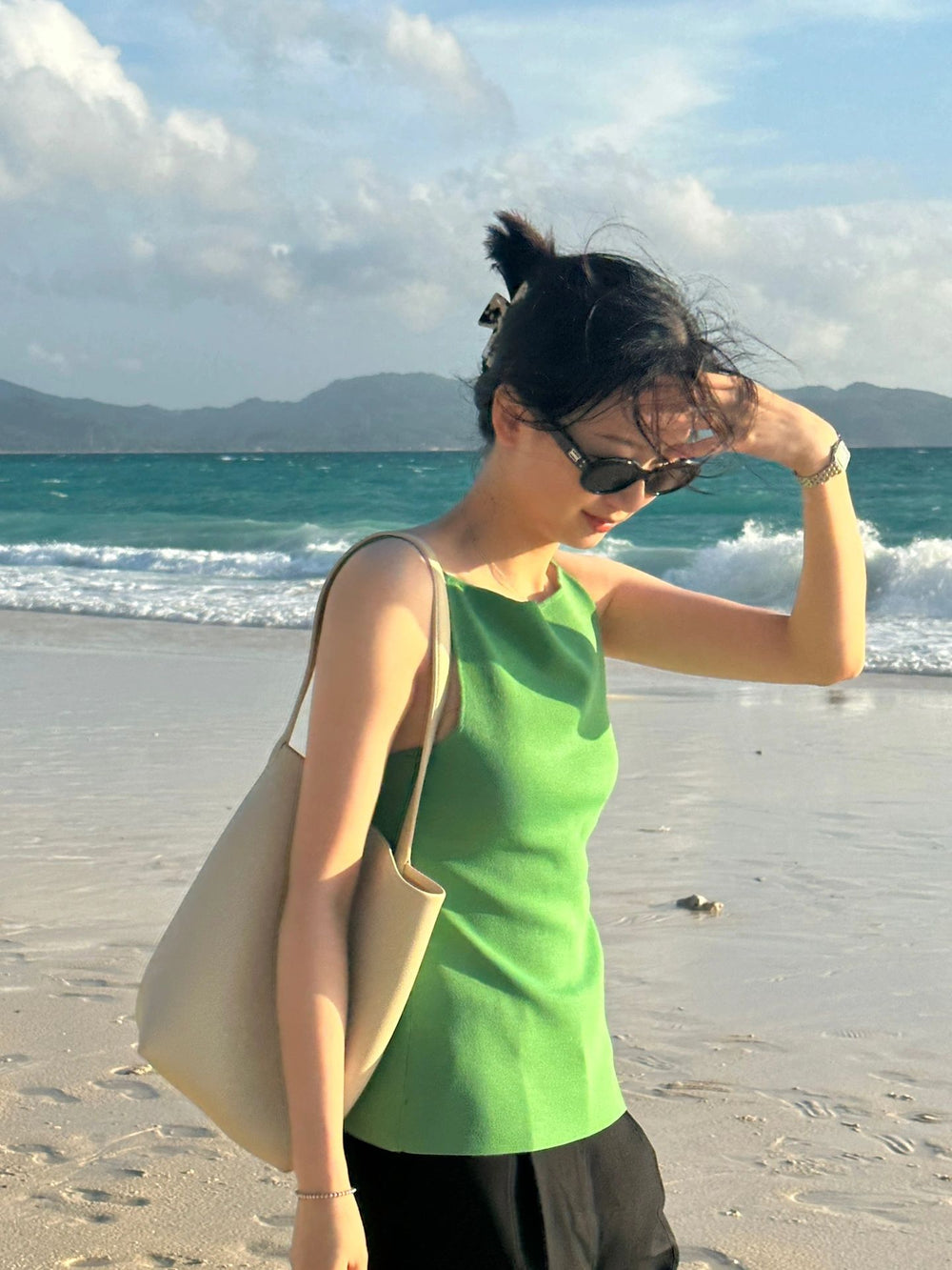 Radiating grace and style, a woman wearing a stunning green top and fashionable sunglasses leisurely walks along the serene beach, enjoying the tranquil surroundings.