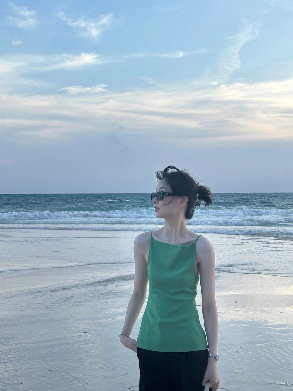 A stylish woman in an elegant green top gracefully poses on the sandy shores of a serene beach wearing her trendy sunglasses.