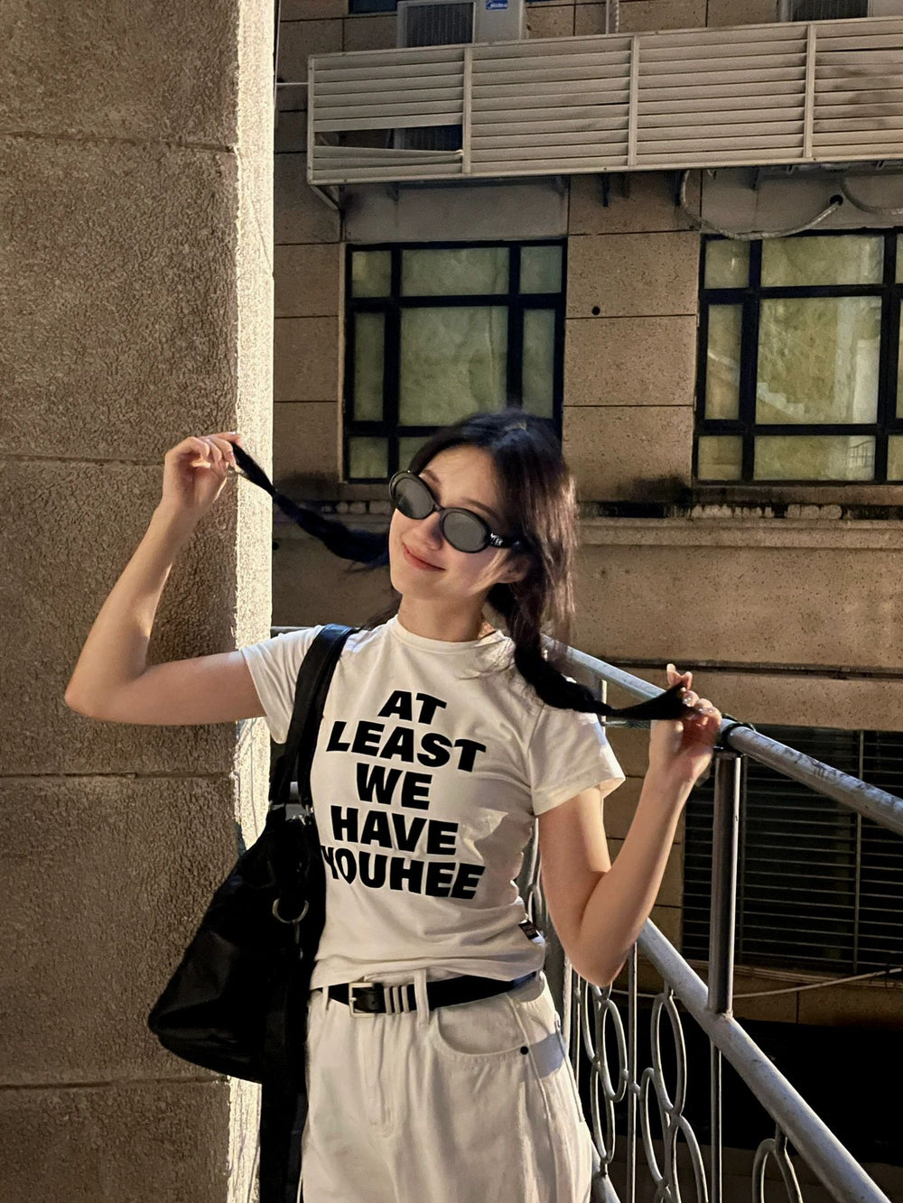 A graceful woman in a white shirt and black skirt standing on a balcony taking in the stunning cityscape wearing her Korean fashionable sunglasses.