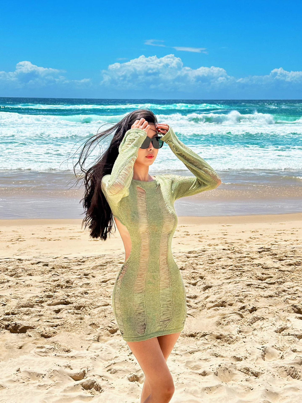 A woman wearing a green dress and korean fashion sunglasses standing on the sandy beach with the waves crashing behind her.