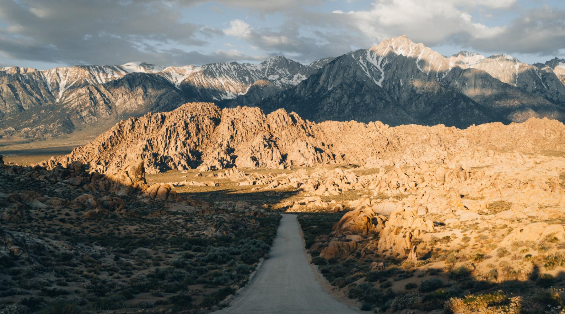 Dirt road leading to Alabama Hills, California.  Sun on snow capped mountains with clouds.