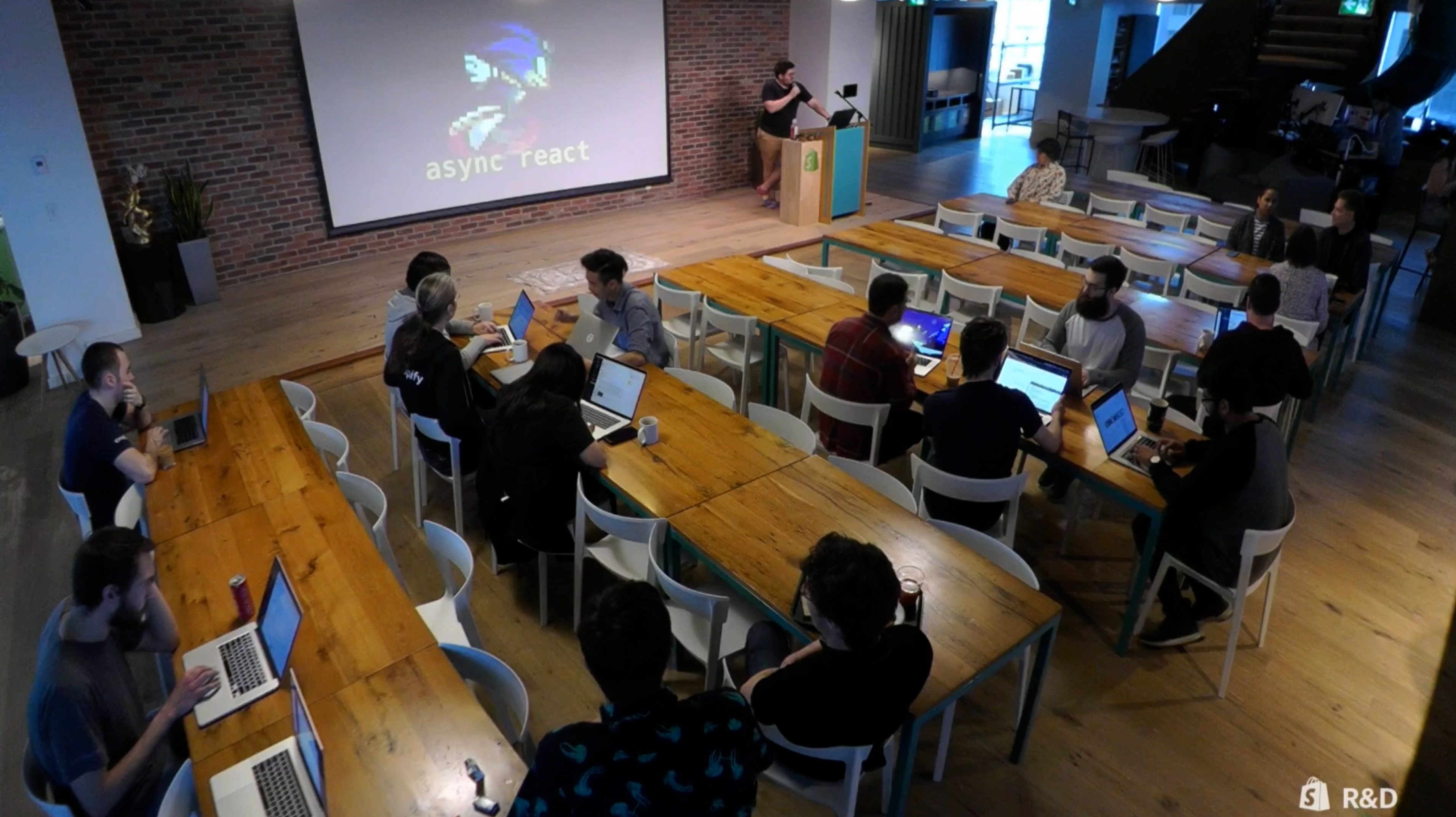 A picture of Shopify employees gathering in the lunch room of the Ottawa Office for Dev Talks.  There are 5 long tables with several people sitting down. The tables are placed in front of a stage with a screen. On that screen is an image of Crash Bandicoot. There is a person standing at a lectern on the stage presenting.