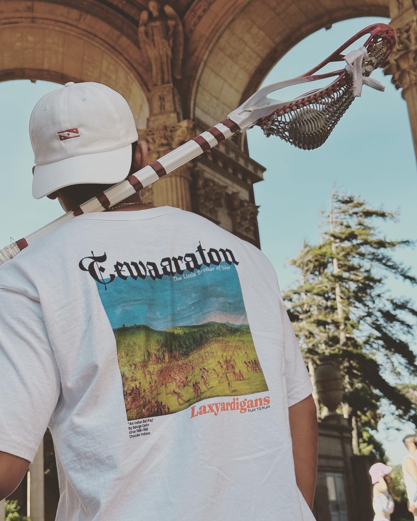 Man wears a hat and shirt by streetwear brand, Laxyardigans, while holding a lacrosse stick, at the Palace of Fine Arts in San Francisco