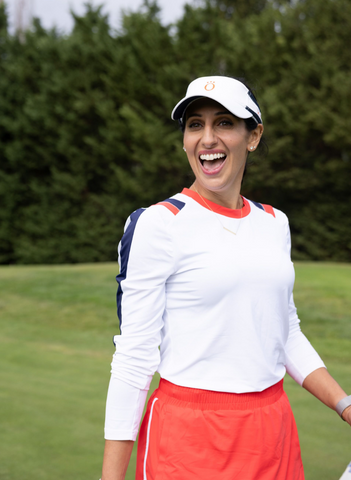 A woman wearing a red skort, white long sleeve shirt, and white visor