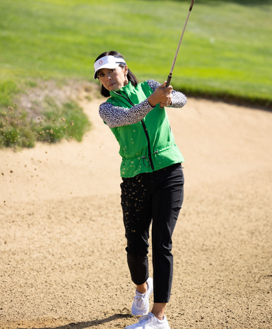 A woman wearing a green vest and black pants, swinging a golf club