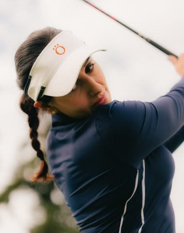 A woman wearing a white visor and blue long sleeved shirt, swinging a golf club