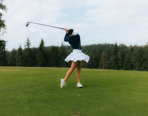 A woman wearing a white skirt and a blue long sleeved shirt, swinging a golf club