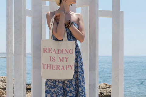 Accessories, bags, gifts. Woman standing by the beach holding a tote bag which reads, reading is my therapy.