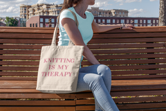 Woman sitting on a park bench with a canvas bag which reads, Knitting Is My Therapy.