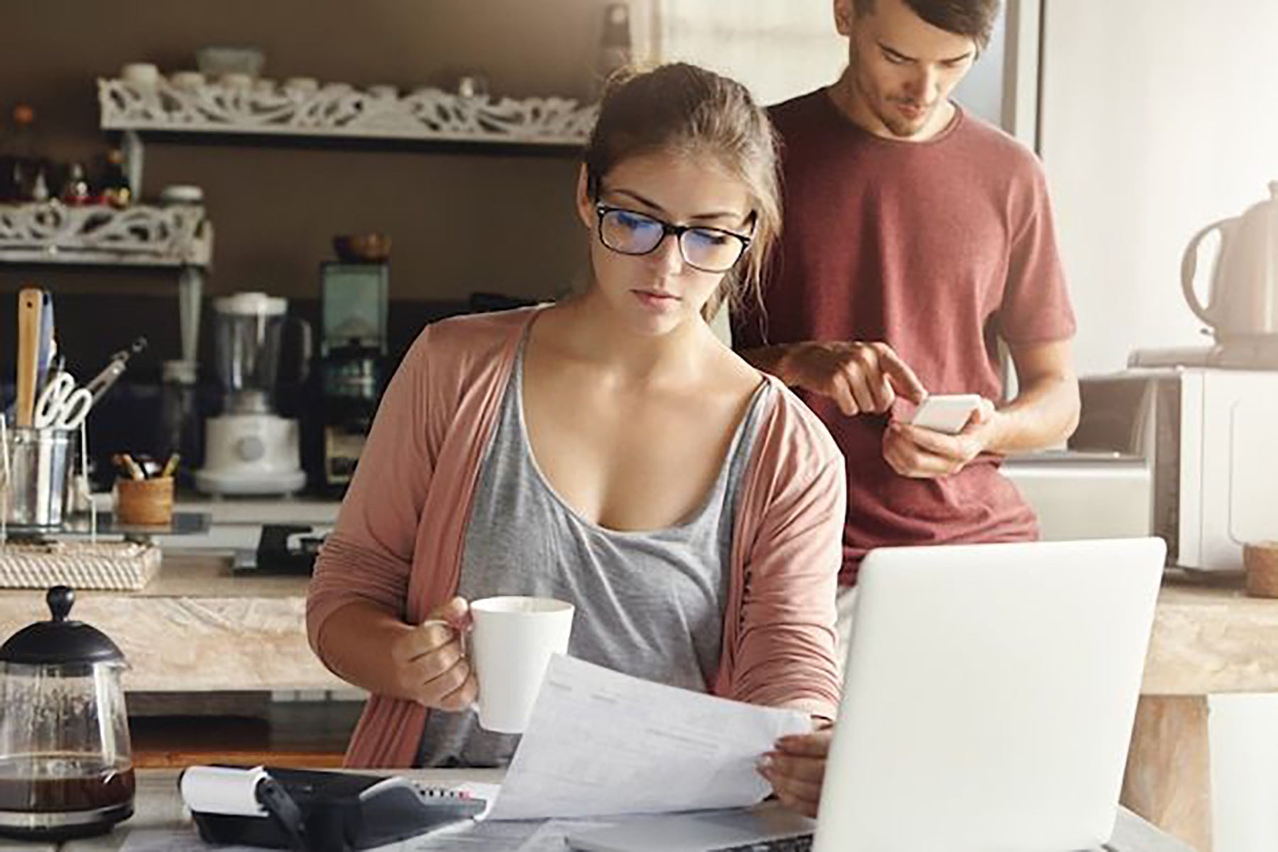 beautiful-serious-young-woman-rectangular-glasses-having-coffee-studying-bill-her-hand-sitting-modern-kitchen-interior-front-open-laptop_273609-1678