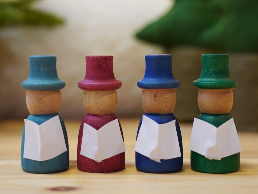 Four peg people with coloured hats hold tiny hymn books with felt trees in the background. Wassailing plays an important part of the 12 days of yuletide celebrations