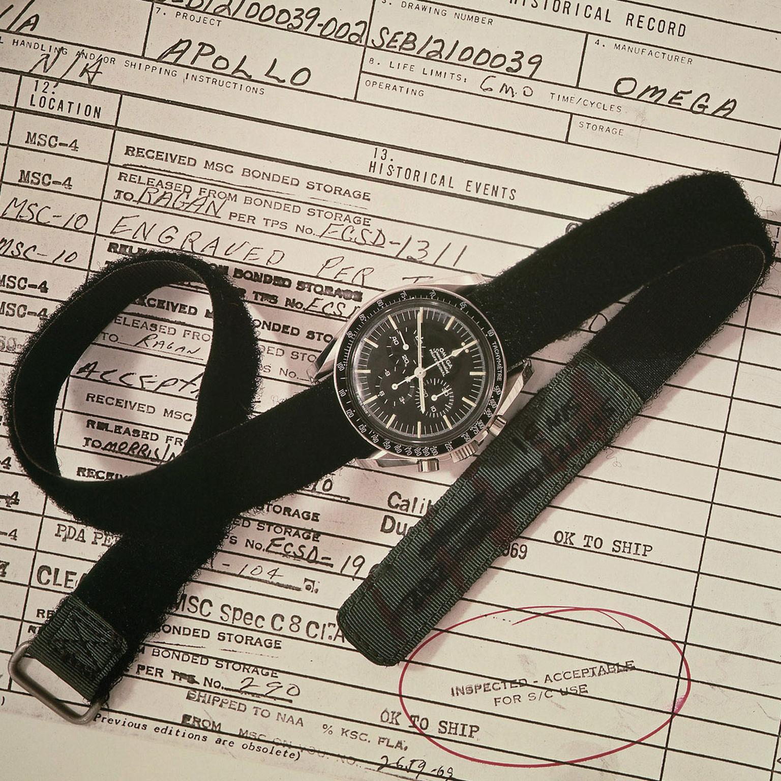 An Omega Speedmaster watch with a black dial, tachymeter bezel, and Velcro strap placed on top of a historical NASA document related to the Apollo program. The document contains handwritten notes and official stamps, including an 'INSPECTED – ACCEPTABLE FOR USE' marking, signifying the Speedmaster’s qualification for space missions. The watch is positioned prominently, emphasizing its association with NASA and space exploration.