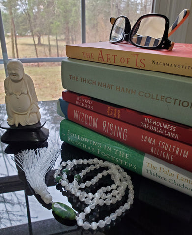 stack of books with a pair of reading glasses on the top and a mala on a glass desk top, a smiling Buddha statue sits off to the left