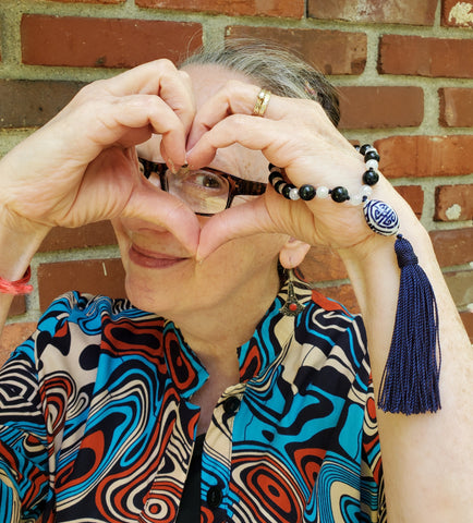 Photo of myself standing in front of a brick wall in a colorful dress. I am peeking through the heart shape with my hands and wearing a quarter mala on my left wrist. 