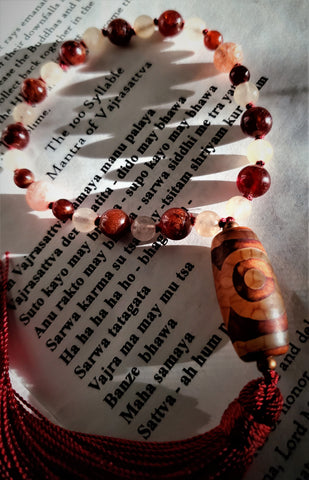 Close up view of Vajrasattva quarter mala with 100 syllable mantra as background. Pink Sunstone and deep red Hessionite Garnet beads with Tibetan agate guru and maroon tassel. Om Vajrasattva Hum.