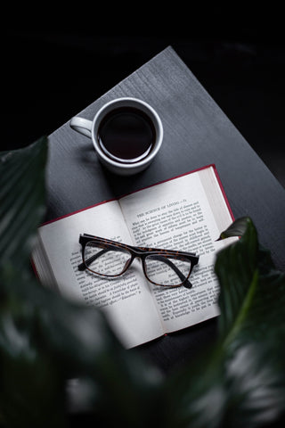 Book opened on desk with a cup of hot coffee beside it and reading glasses placed on top of book.