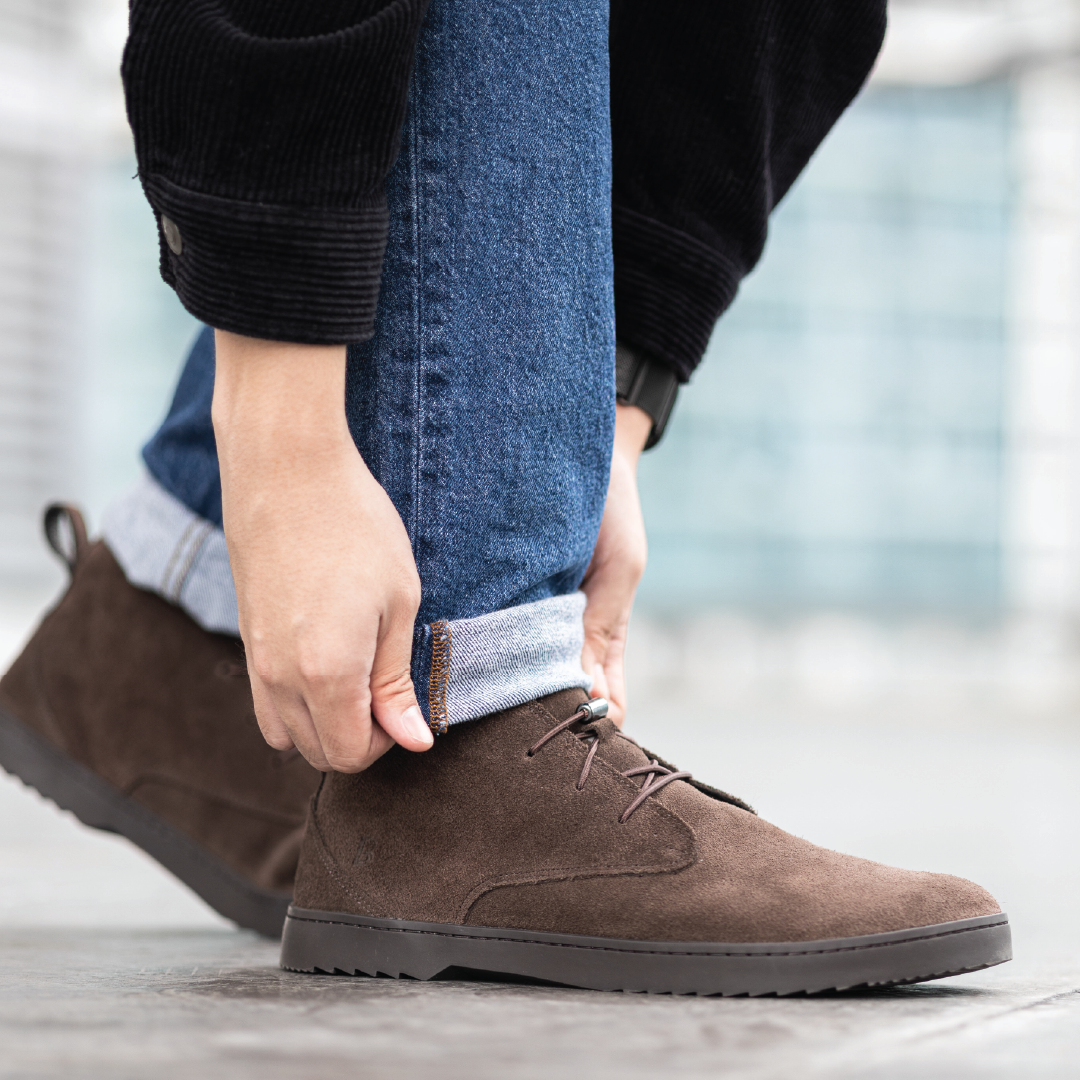 Person adjusting jeans while wearing brown suede shoes.