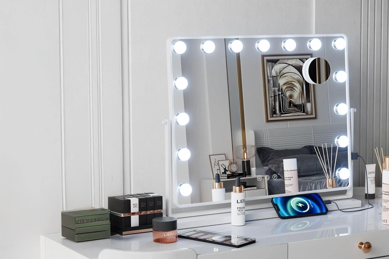 Une vue frontale d'un grand miroir de maquillage lumineux de style "Hollywood", avec 15 ampoules LED rondes encastrées le long de son cadre blanc. Le miroir mesure 90 x 60 cm comme indiqué dans le texte superposé. Il est posé sur une coiffeuse blanche où plusieurs produits de beauté et accessoires sont disposés, incluant un smartphone en charge par induction, des flacons et des boîtes. Le miroir reflète partiellement une chambre avec un lit et un tableau sur le mur. En haut à gauche, un texte indique "Hollywood - Miroir LED - 90 x 60 cm".