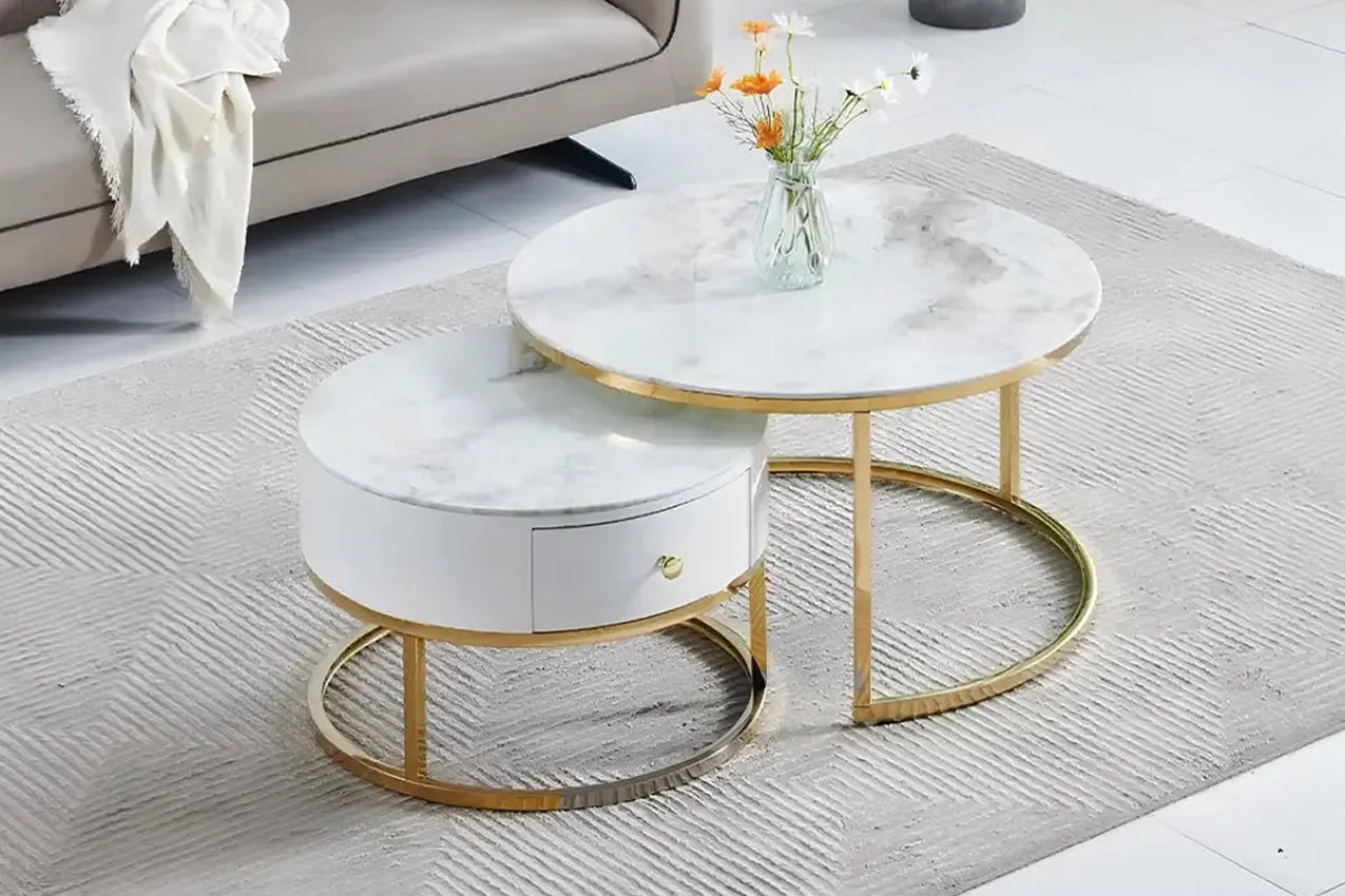 Deux tables basses rondes gigognes avec plateaux en marbre blanc et bases dorées, l'une plus grande et l'autre plus petite avec un tiroir blanc, posées sur un tapis gris clair devant un canapé beige.