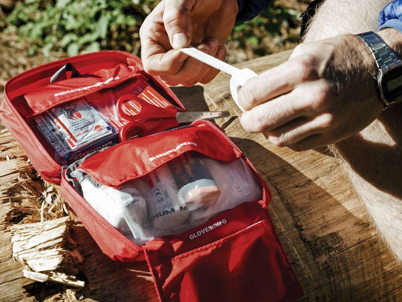 A person applies medical tape from a well-stocked red Lifesystems first aid kit, placed on a log in a forest setting.
